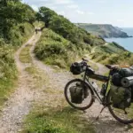 A robustly loaded touring bicycle pauses off-centre at a fork in a sun-dappled coastal cycle path